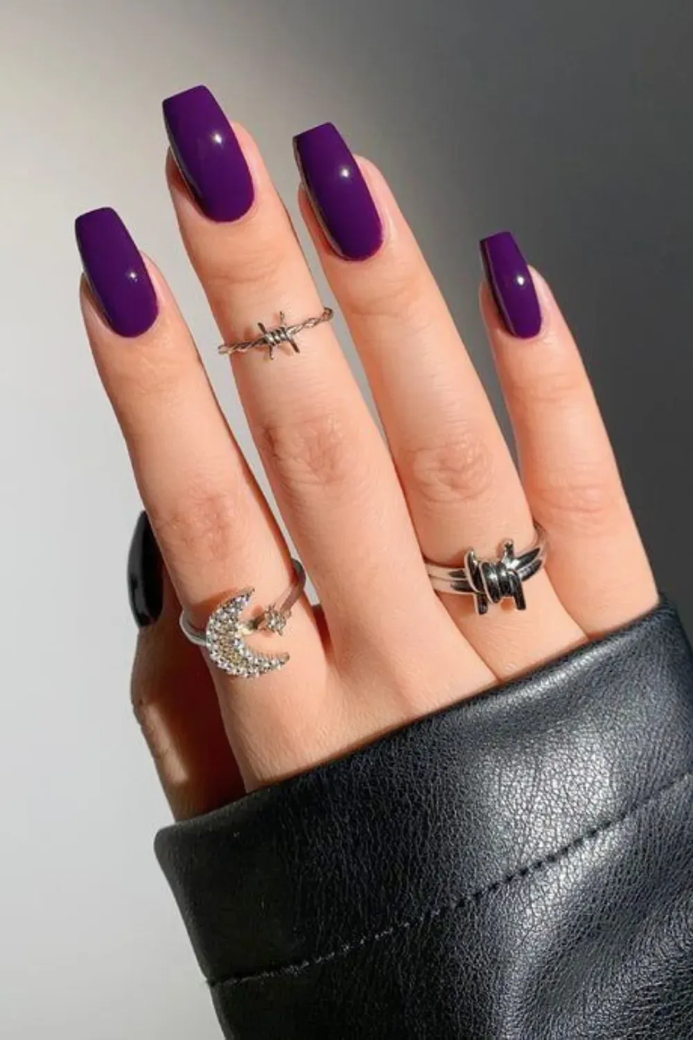 A hand featuring square-shaped purple nails adorned with silver rings, showcasing a chic and elegant style.