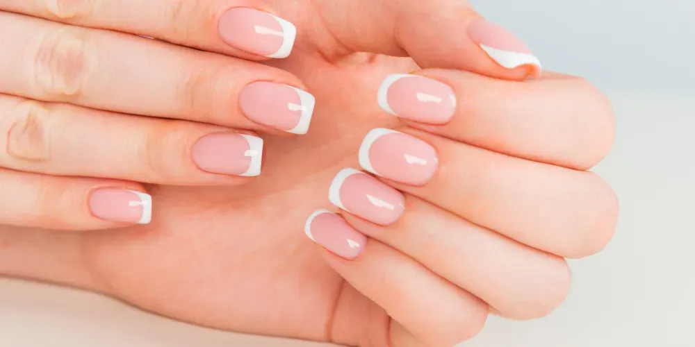 A close-up of hands with well-manicured nails featuring a French tip design. The nails are painted in a soft pink shade with a clean white tip, giving a classic and elegant appearance