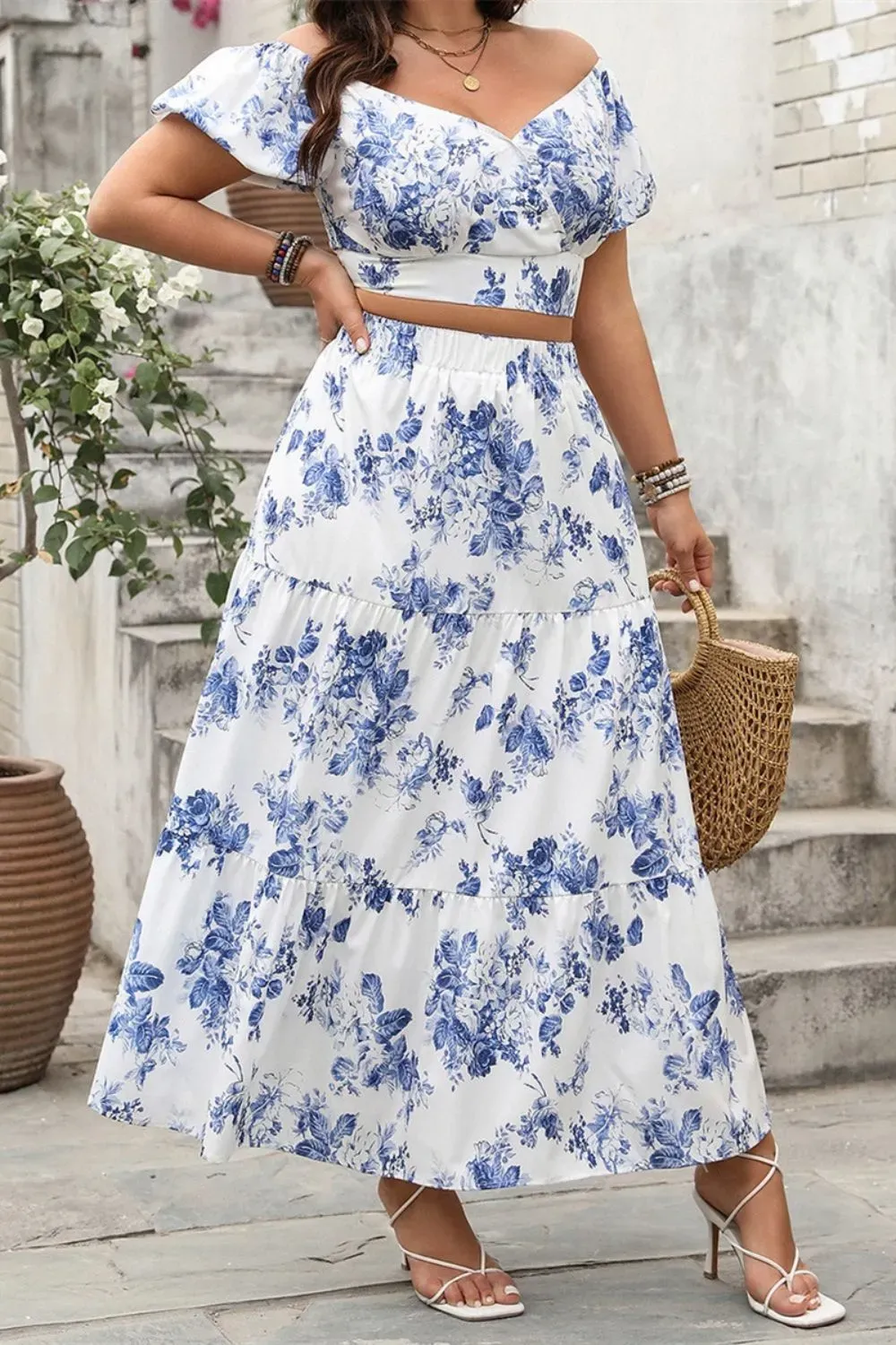 Woman in blue and white floral off-shoulder crop top and tiered maxi skirt set with white heels and a woven handbag.
