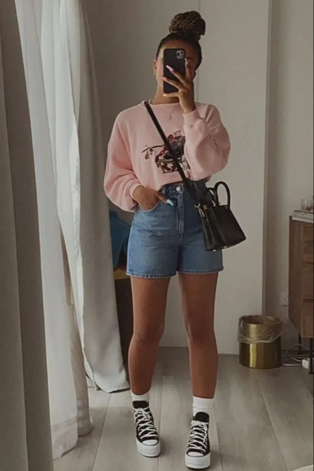 A woman wearing a casual T-shirt and a denim short, holding a black handbag in front of a mirror