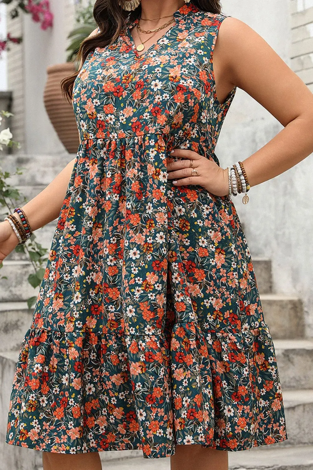 Woman in green floral sleeveless dress with ruffled hem and layered jewelry standing on stone steps.