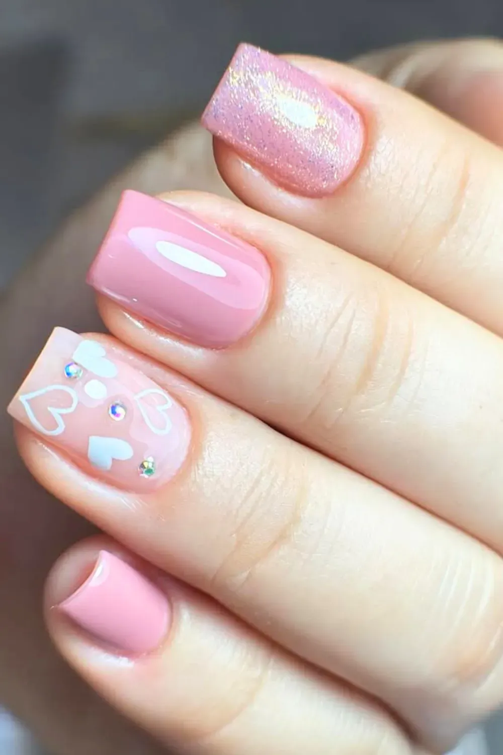 A hand with minimalistic pink nails adorned with heart designs and gemstones.