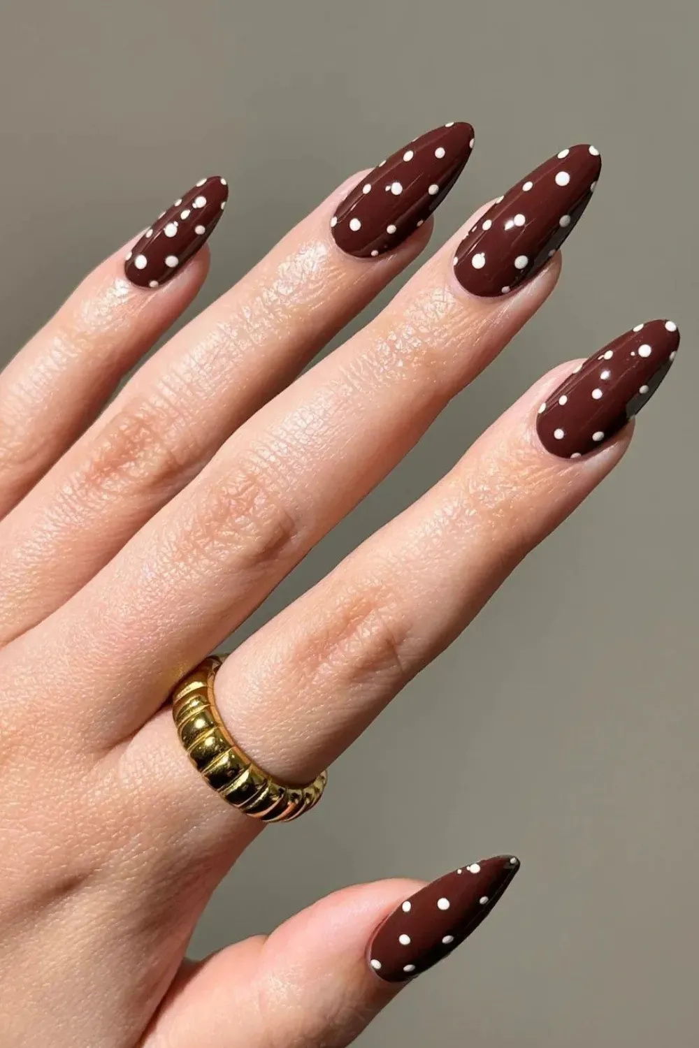 Brown nails with white polka dots on almond-shaped tips, paired with a gold ring, giving a playful and elegant fall manicure look
