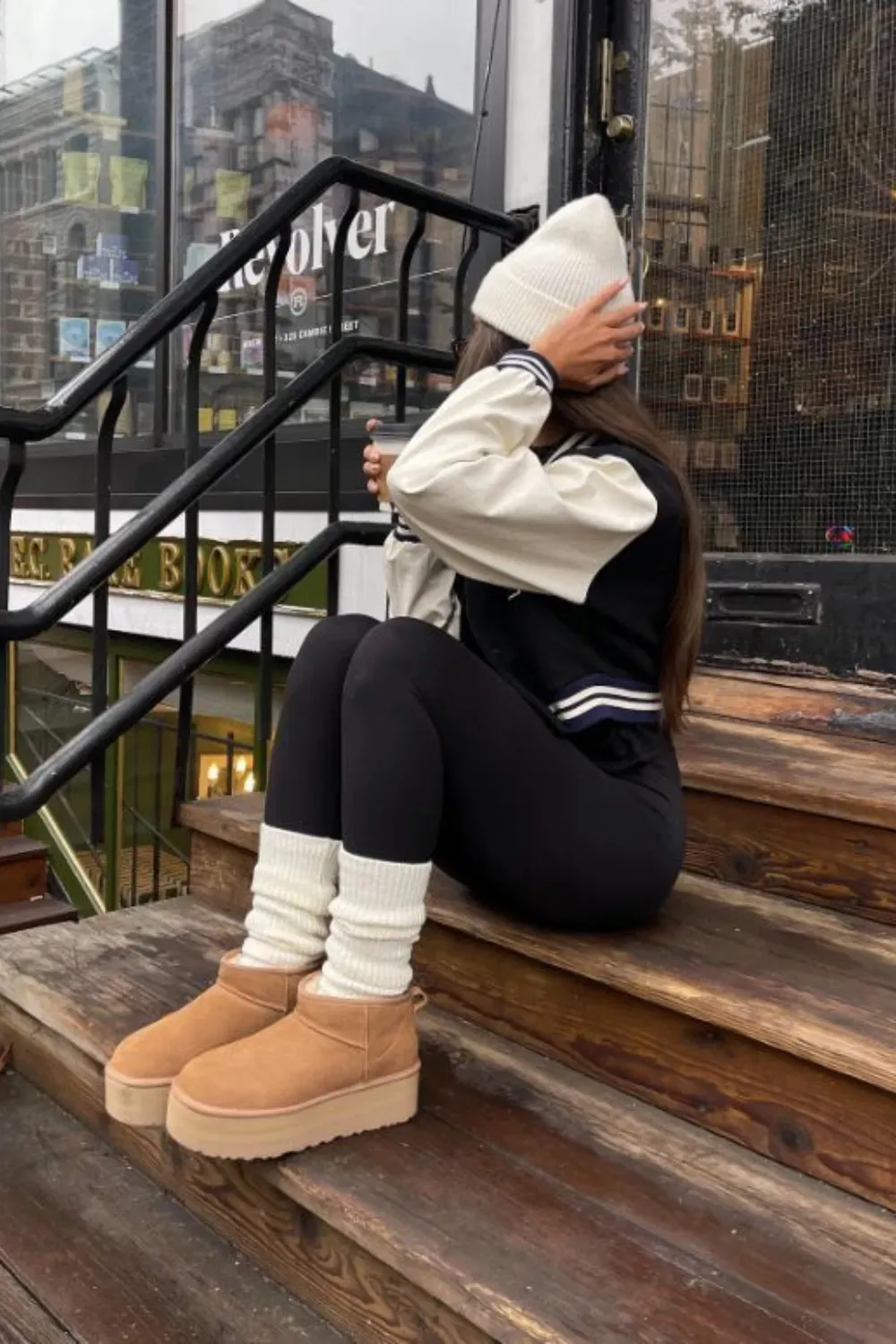 A woman sits on wooden stairs, wearing a white beanie and a black and white varsity jacket. She has on black leggings, white leg warmers, and tan UGG Classic Mini Platform boots. She is holding a coffee cup