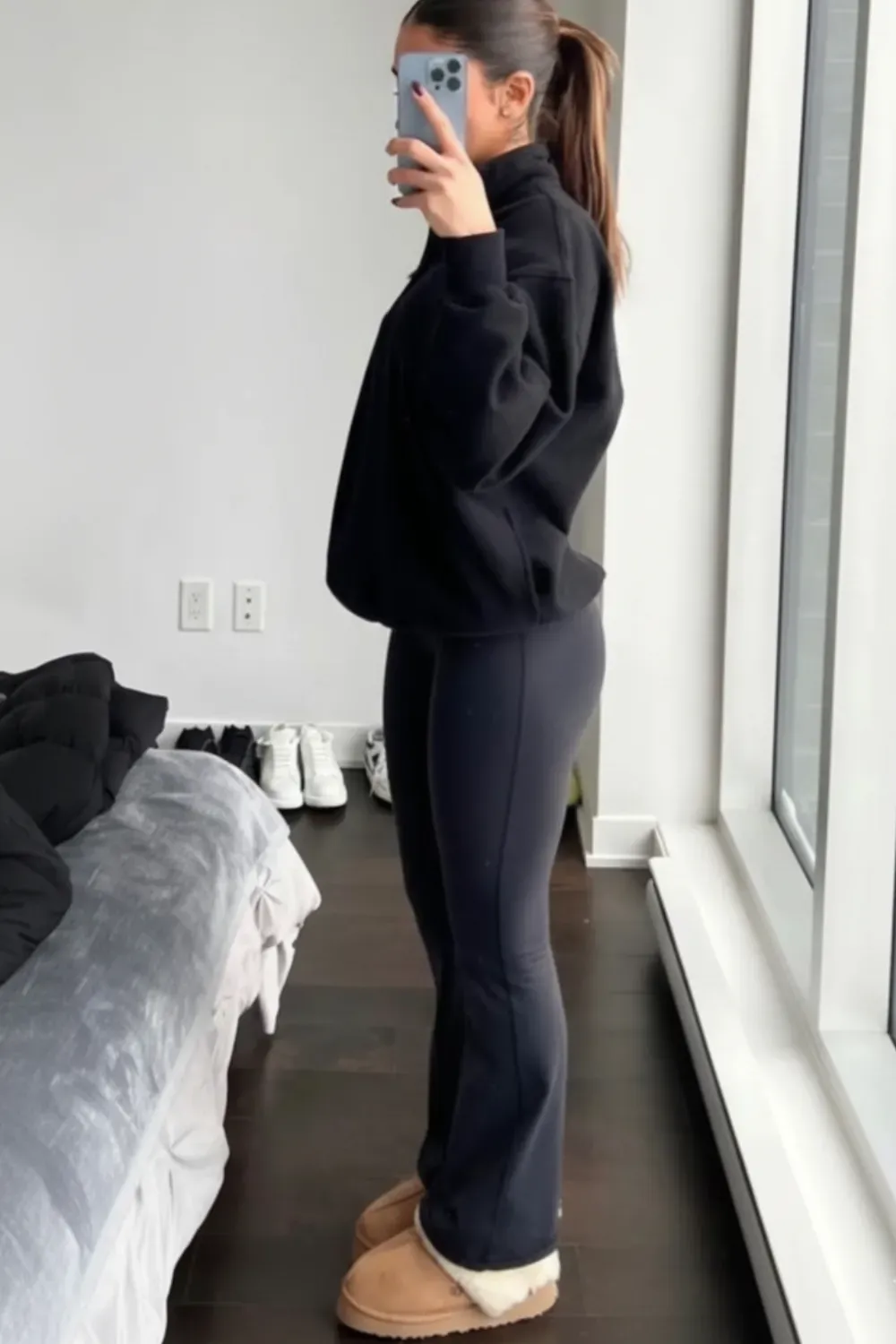A woman is shown in a side profile, taking a mirror selfie. She is wearing a black high-collared sweatshirt, black flare leggings, and tan UGG Classic Mini boots