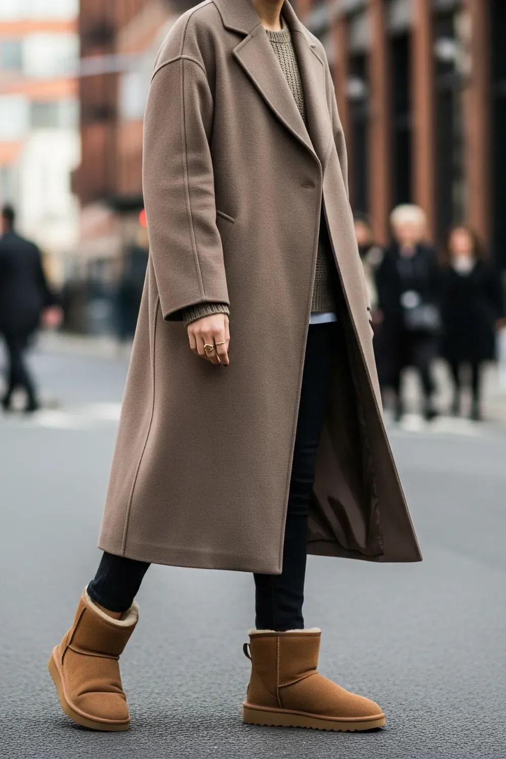 A woman is walking on a city street, wearing a long brown overcoat, black jeans, and tan UGG Classic Tall boots. She has on a ring and a watch