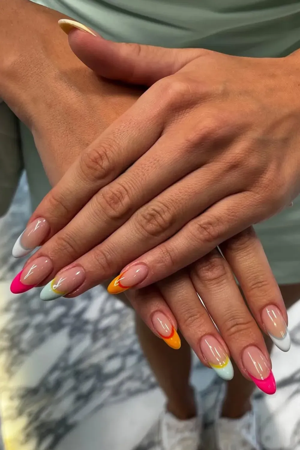 A pair of hands showcasing vibrant, multi-colored French tips in neon shades of pink, orange, yellow, and green, with a smooth nude base