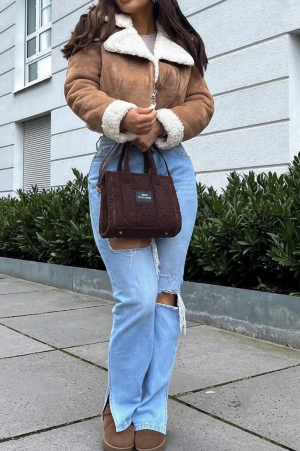 Cozy Shearling Jacket with Ripped Jeans - A woman wearing a brown shearling coat, ripped blue jeans, brown boots, and carrying a small handbag, standing outdoors.