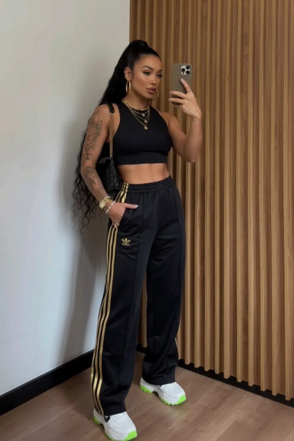 Effortless Casual in Adidas Tracks - A woman wearing a black crop top, Adidas track pants with gold stripes, and white sneakers with green soles. She accessorizes with gold necklaces and earrings, posing casually in front of a wooden background