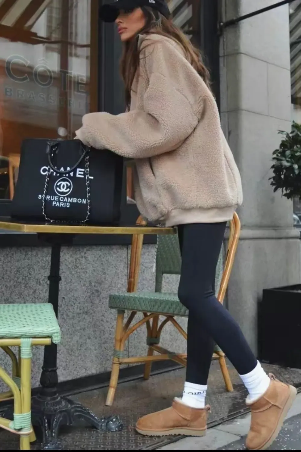 A woman in a black baseball cap and a cream-colored sherpa hoodie stands at an outdoor cafe table. She is holding a black Chanel tote bag and wearing black leggings, white socks, and tan UGG Classic Mini boots.