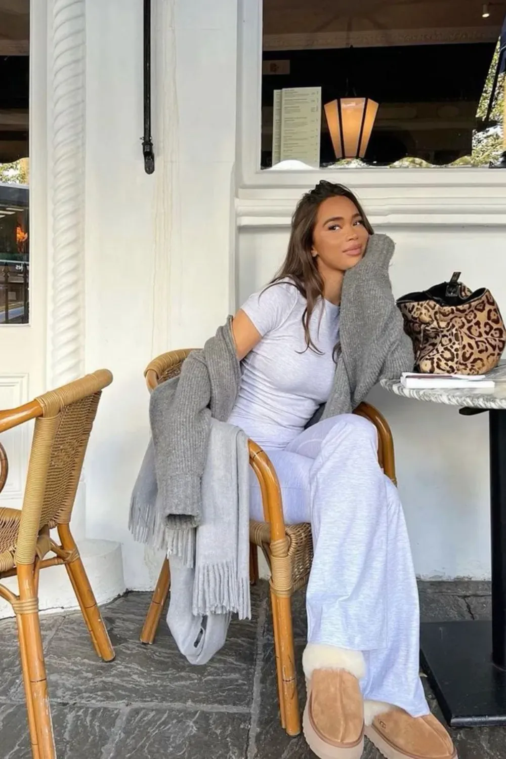 A woman sits at an outdoor cafe table, wearing a light grey matching long-sleeve top and wide-leg trouser set. She has a leopard print bag on the table and is wearing tan UGG Tasman slippers.