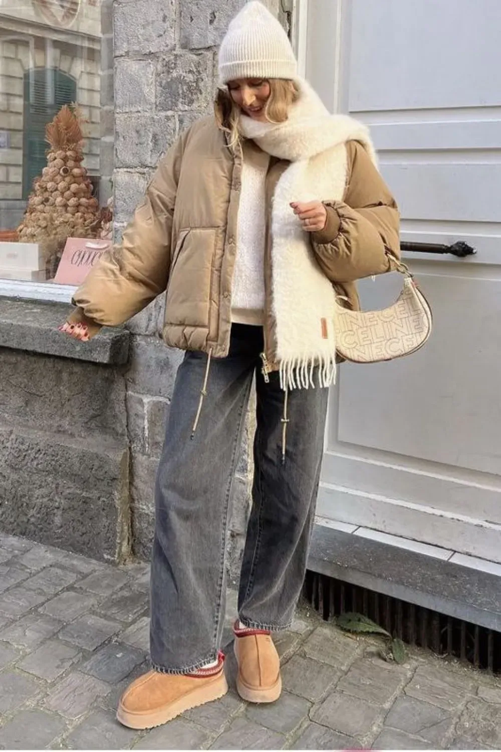 A woman is standing outdoors, wearing a white beanie, a thick white scarf, and a beige puffer jacket. She has on dark grey wide-leg jeans with red-striped socks and tan UGG Tazz slippers. She is holding a cream-colored handbag with a logo