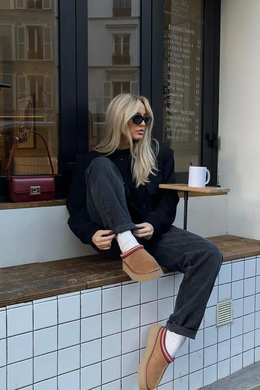 A blonde woman with sunglasses is sitting on a tiled outdoor bench next to a cafe. She is wearing a black jacket and dark grey, wide-leg jeans with red-striped socks and tan UGG Tazz slipper