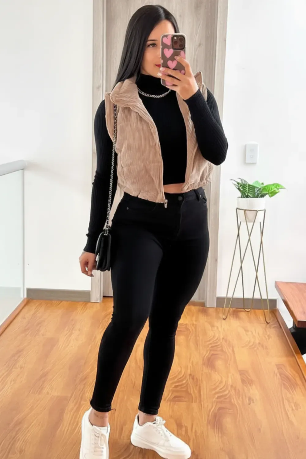  Casual Vest and Black Top Fall Outfit - A woman in a corduroy vest, black top, skinny black jeans, and white sneakers, standing in a modern interior.