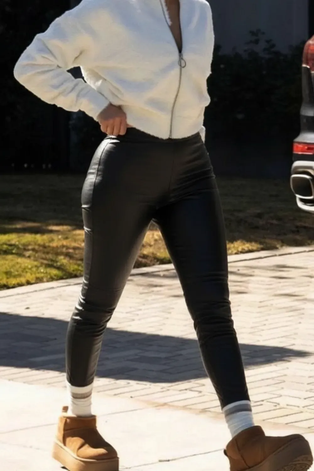 A woman is walking outdoors in a pair of shiny black leather trousers and a cropped white zip-up jacket. She is also wearing white socks and brown UGG Classic Mini Platform boots