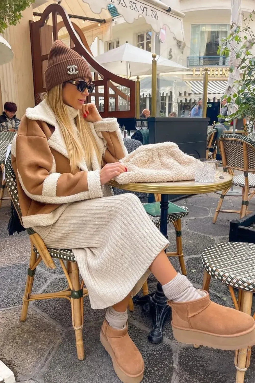 A woman is sitting at an outdoor cafe table in Paris. She is wearing a brown beanie with a white Chanel logo, sunglasses, and a brown and cream shearling jacket. She has on a ribbed cream dress, tan socks, and UGG Classic Mini Platform boots