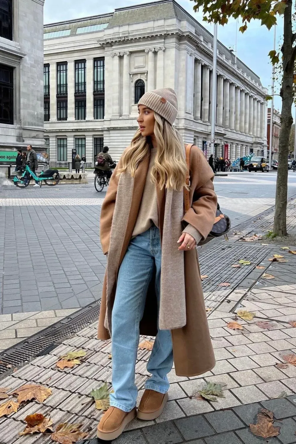  A woman stands on a leaf-strewn city sidewalk on a cloudy day. She is wearing a neutral-colored beanie, a long camel coat, and a matching scarf. She has on light-wash blue jeans and tan UGG Tazz slippers