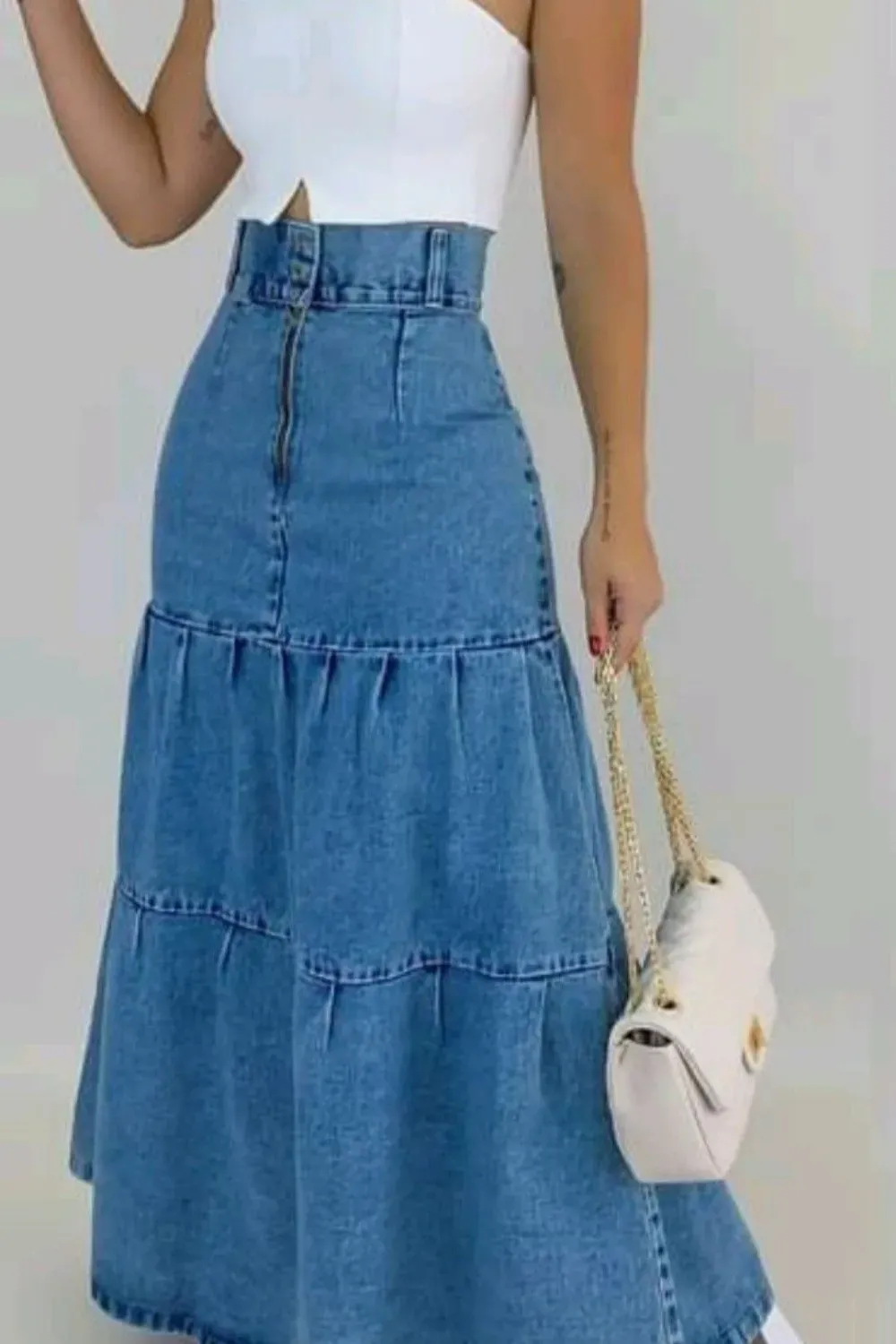 Tiered Denim Maxi Skirt with White Crop Top	Woman wearing a high-waisted, tiered, light blue denim maxi skirt with a white square-neck crop tank top, holding a white quilted bag.