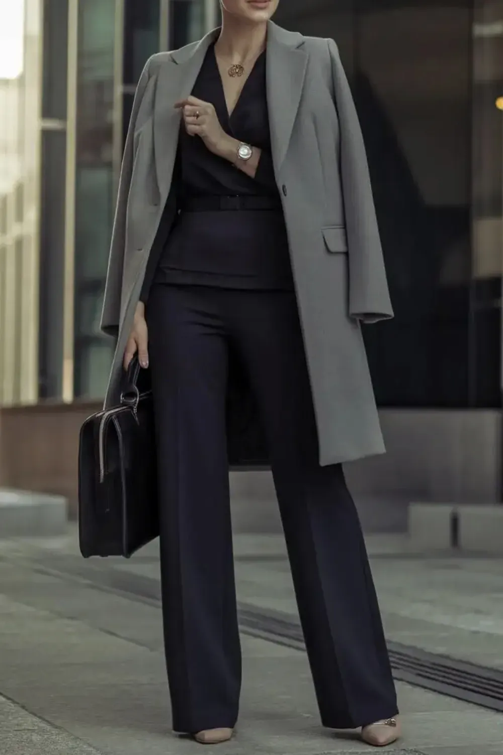 Monochromatic Power Suit with Grey Coat  - A woman standing outside wearing a black v-neck top, black wide-leg trousers, a black belt, and a light grey long coat draped over her shoulders. She holds a black briefcase.