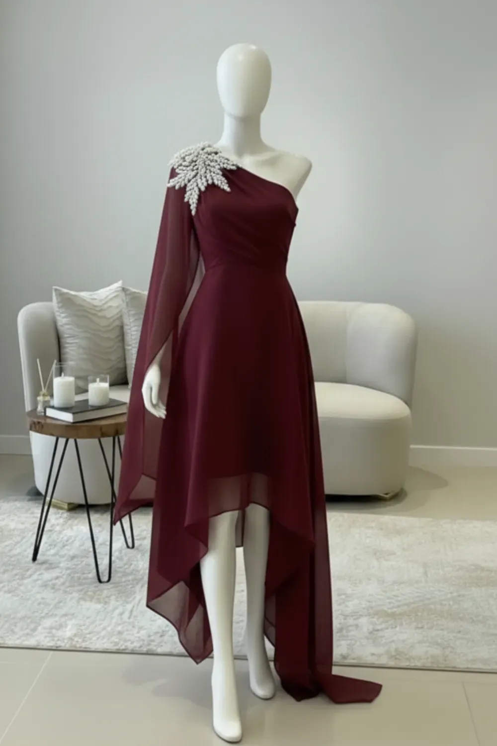 Asymmetrical High-Low Burgundy Chiffon Dress with Pearl Embellishment - Mannequin in a contemporary setting wearing a burgundy high-low dress with a sheer cape-sleeve and a large, intricate pearl embellishment on the single shoulder.