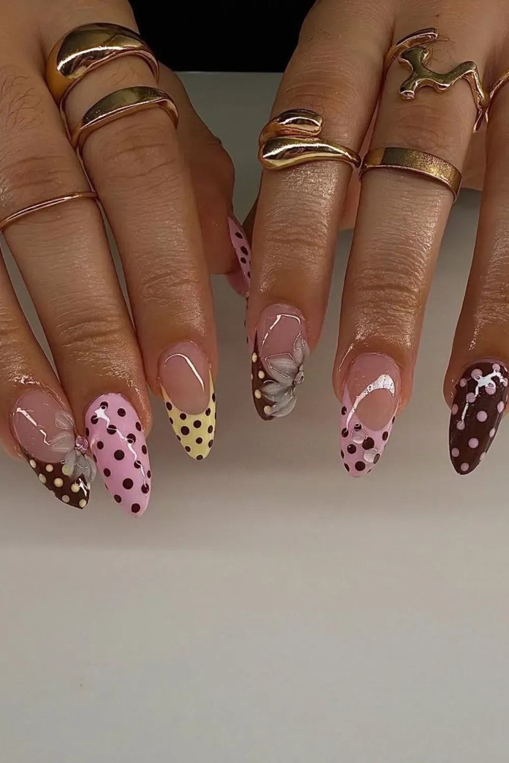 Long almond nails featuring yellow, pink, and brown tips with black and pink polka dots, and a 3D floral embellishment.