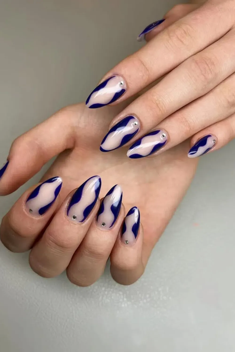 Navy Abstract Wave Nails - Almond-shaped nails with a nude base and glossy, flowing abstract waves in deep navy blue, with small rhinestone accents.
