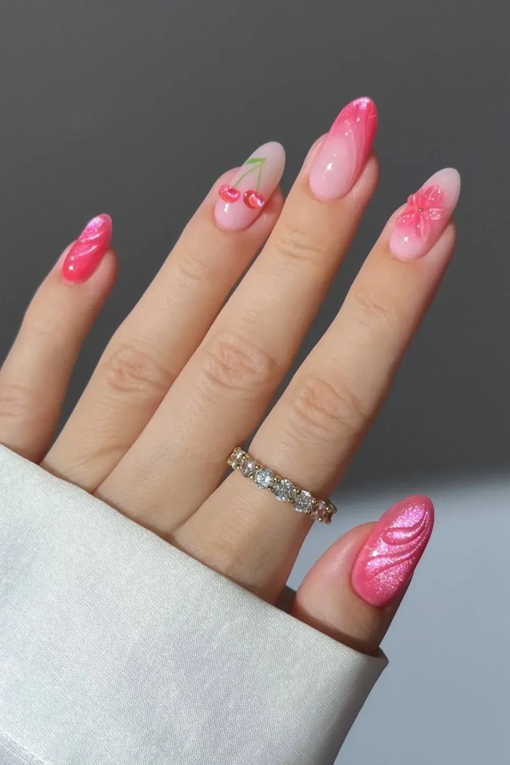 Hand with a mix of pink nail designs, including ombre with cherries, a 3D flower, and metallic pink waves.