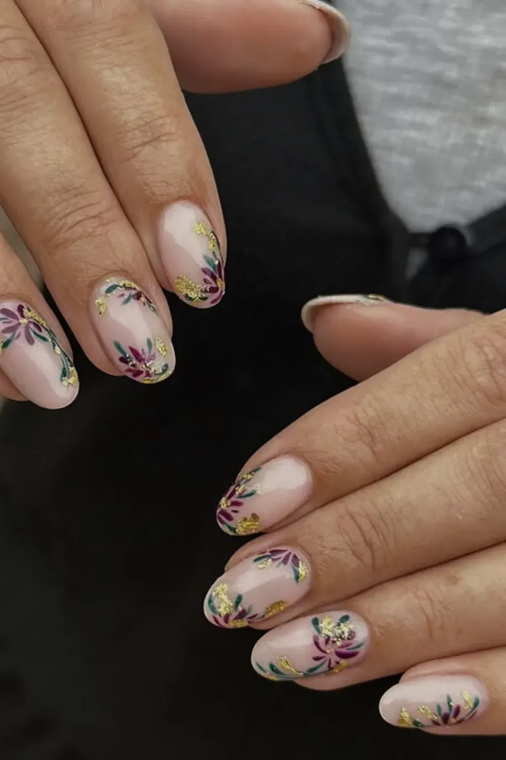 Hands with short oval nude nails featuring abstract floral designs in purple, green, and gold flakes.