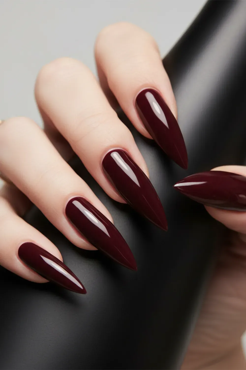 Hand holding a black object, showing very long, glossy stiletto nails in a solid, deep burgundy color.