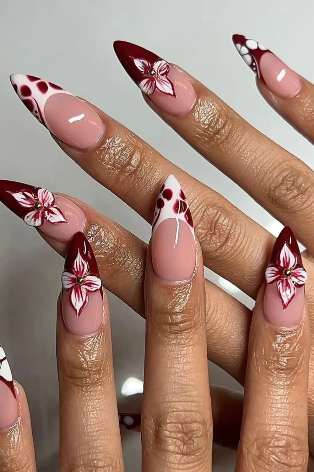 stiletto nails design of nude bases, burgundy French tips, white polka dots on burgundy, and white and red 3D floral accents with gold centers