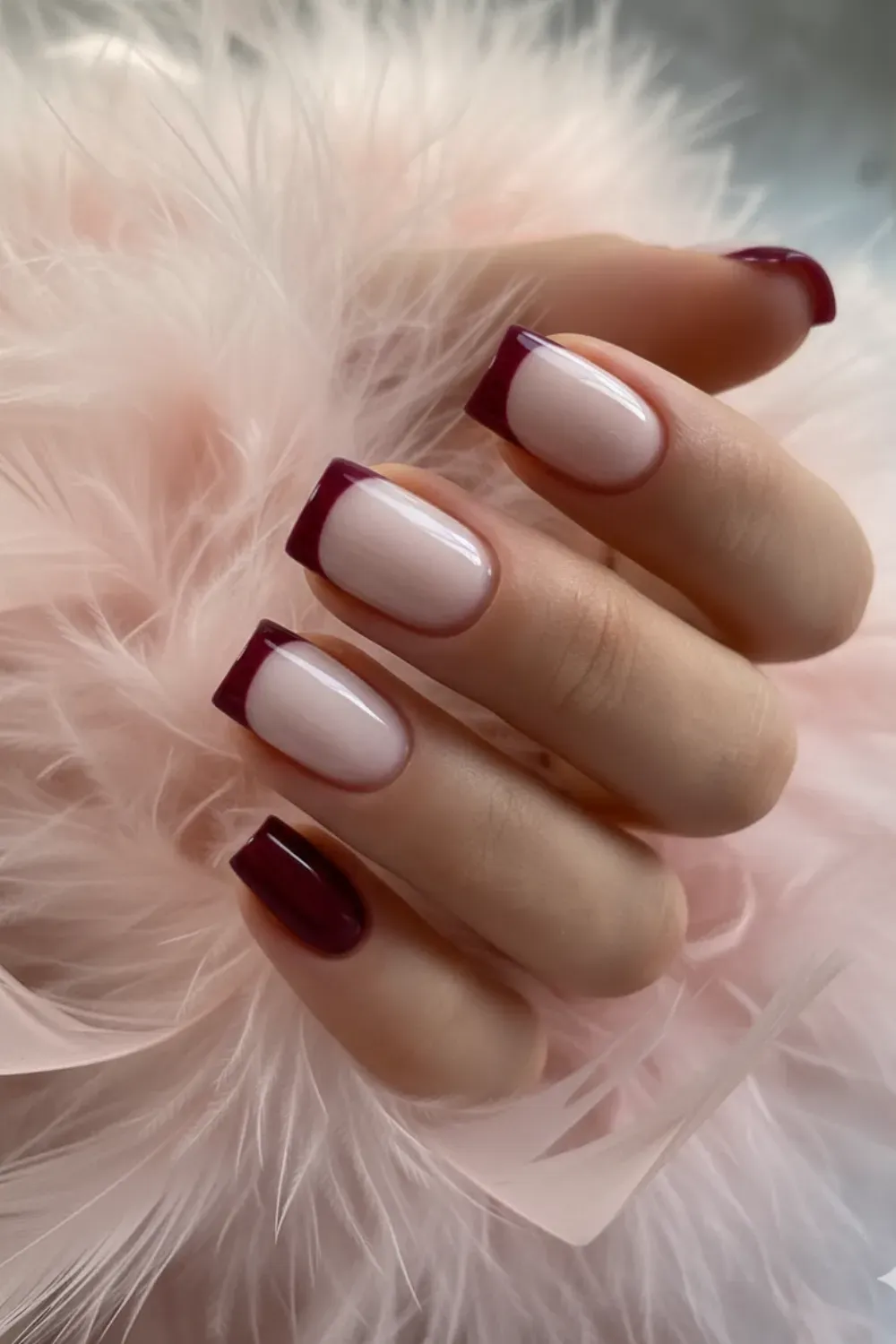 Hand holding a pink feather, showing short square nails with a nude base and distinct burgundy French tips.