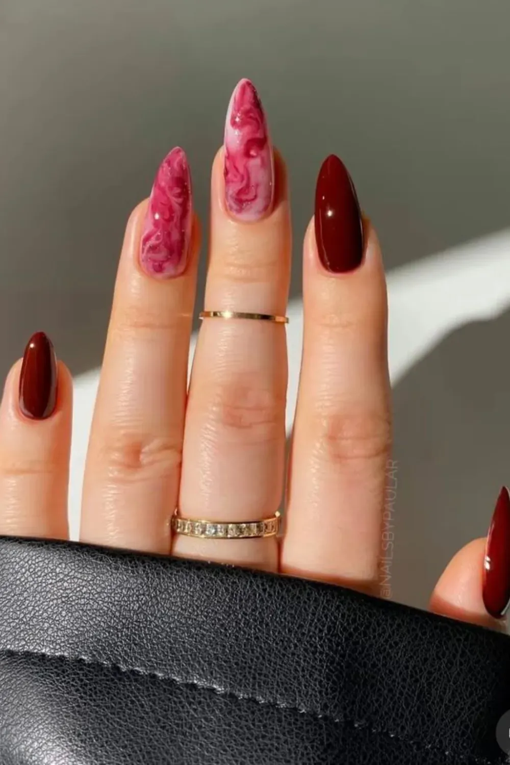 Hand with long almond nails, some in solid glossy burgundy and others with a white and burgundy marble swirl design.
