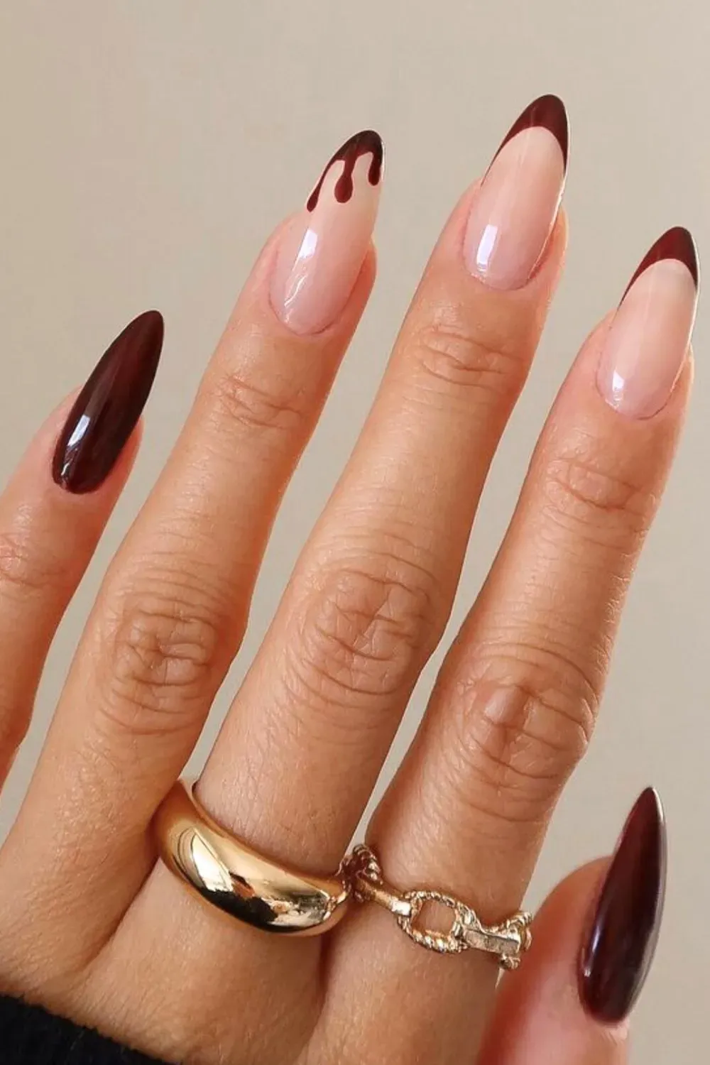 Hand with long almond-shaped nails featuring a nude base and dark brown French tips with a "drip" effect.