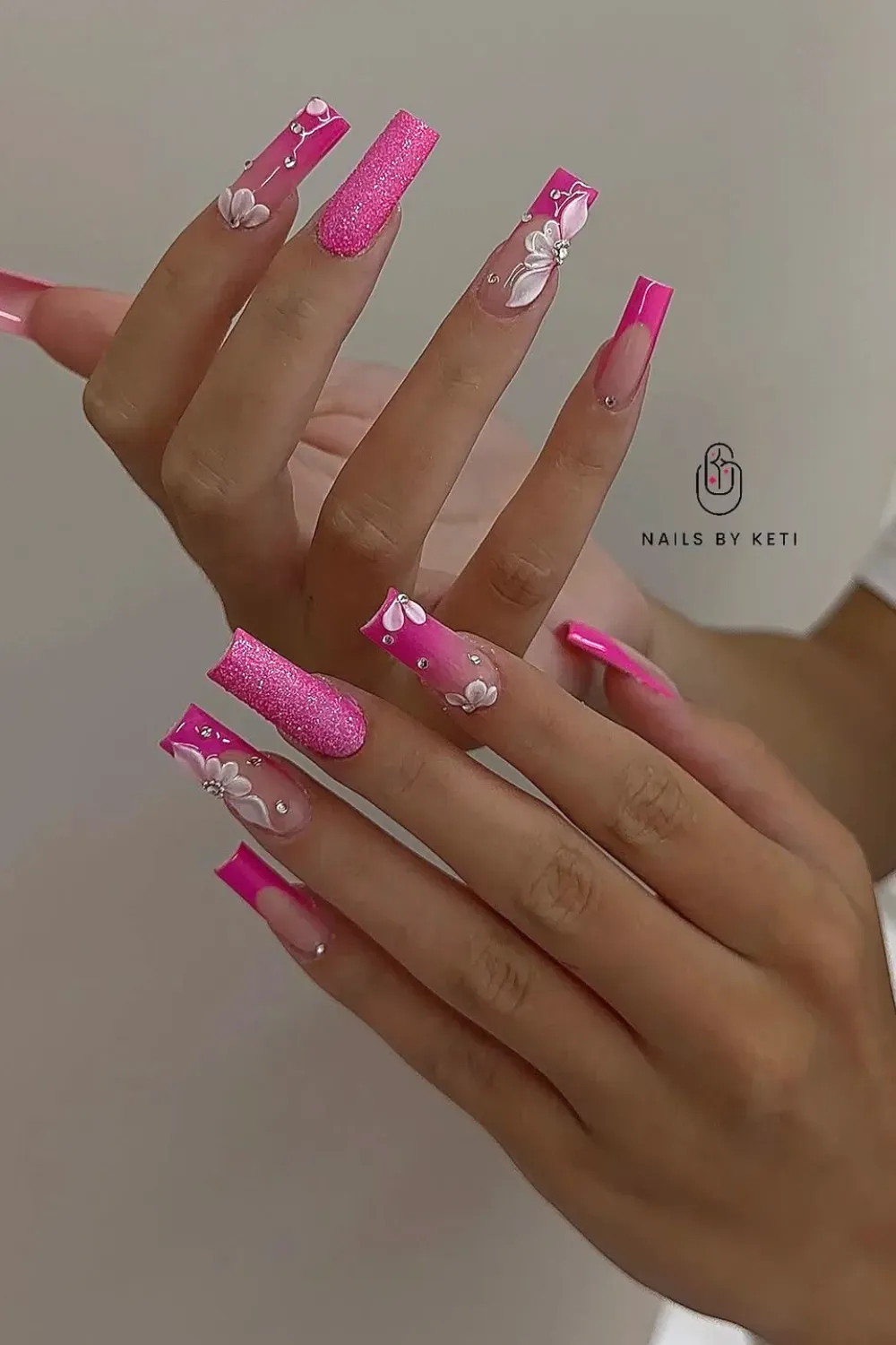 Hands with long coffin-shaped nails, some are hot pink glitter, others have hot pink French tips with white 3D flowers.
