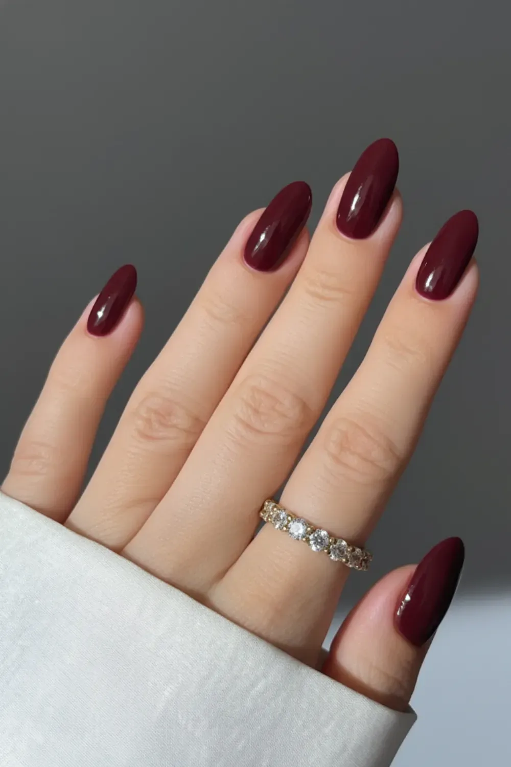 Hand with long, glossy almond-shaped nails painted in a solid, deep burgundy color, wearing a diamond eternity ring.