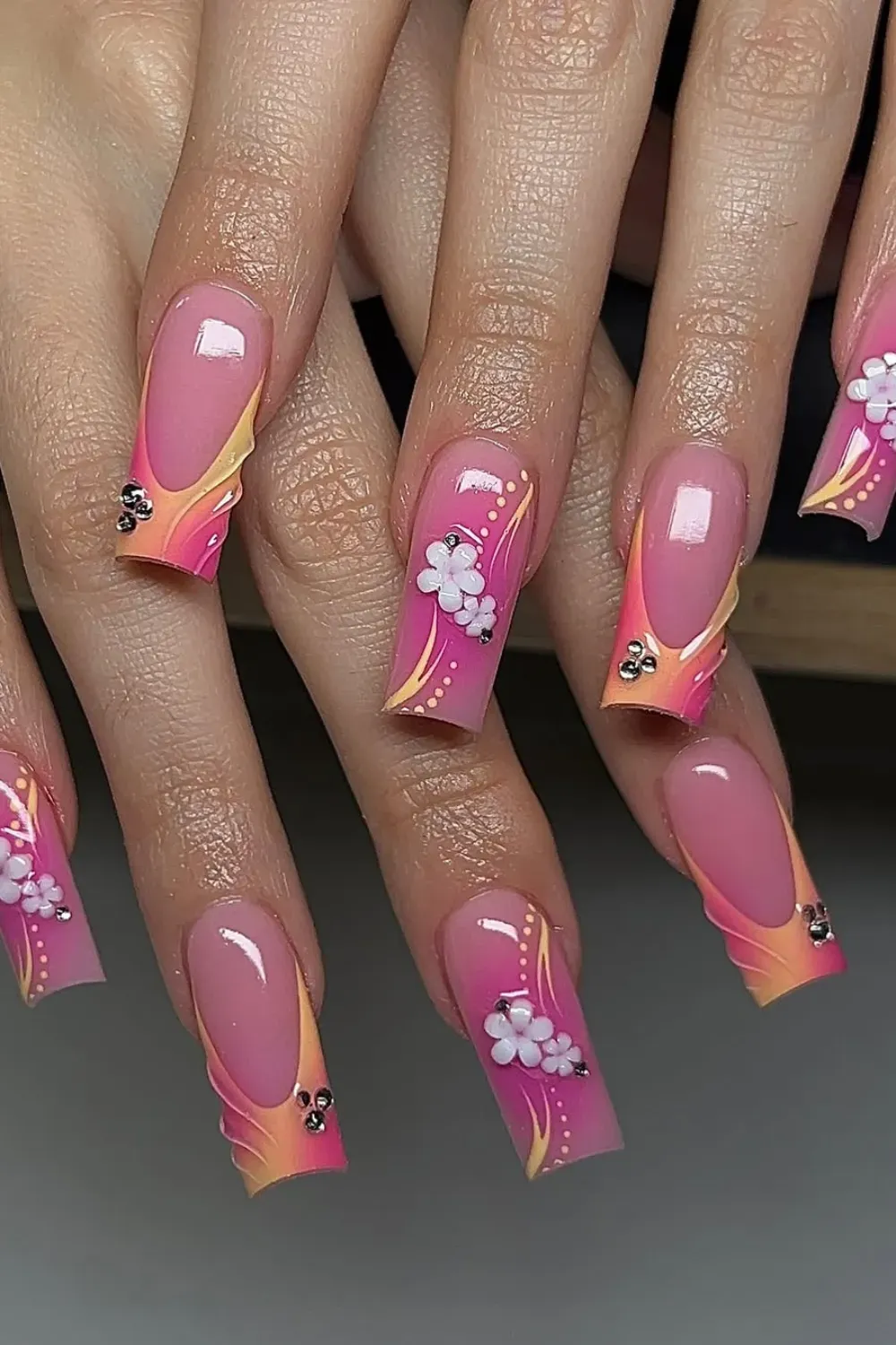 Hands with very long square pink French tip nails, featuring wavy yellow accents, white 3D flowers, and silver studs.