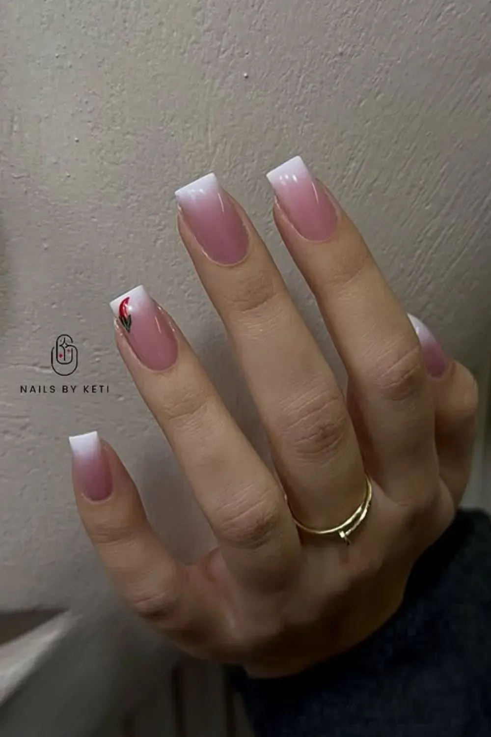 Hand with square-shaped nails featuring a pink to white ombre French fade, with a small red rose design on the ring finger.