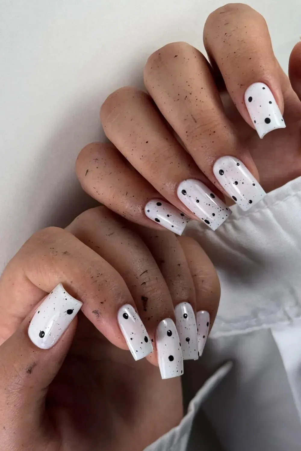 Hands with long square white nails featuring irregular black speckle dots.