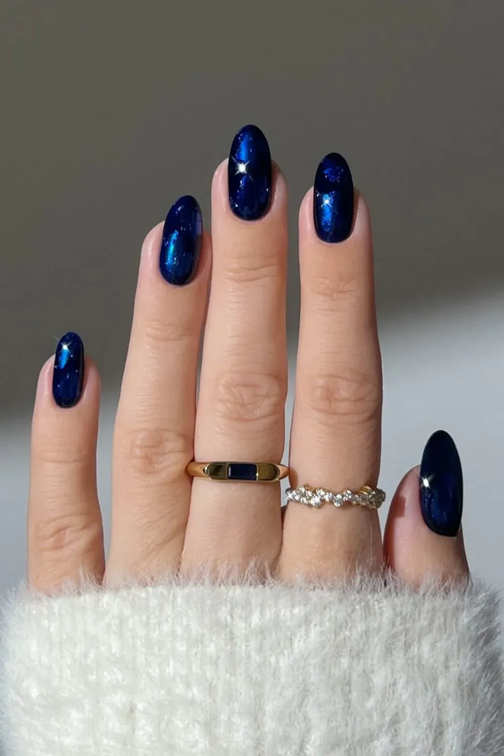 Deep Shimmering Blue Oval Nails with Gold Rings Oval-shaped nails in a deep, shimmering blue polish, worn with gold rings on fingers and a white fluffy sweater cuff.