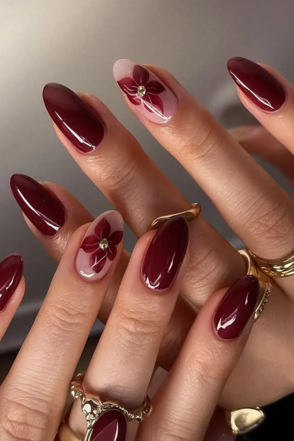 Two hands with oval-shaped glossy burgundy nails, some featuring a light pink base with a burgundy floral design and a gold bead center.
