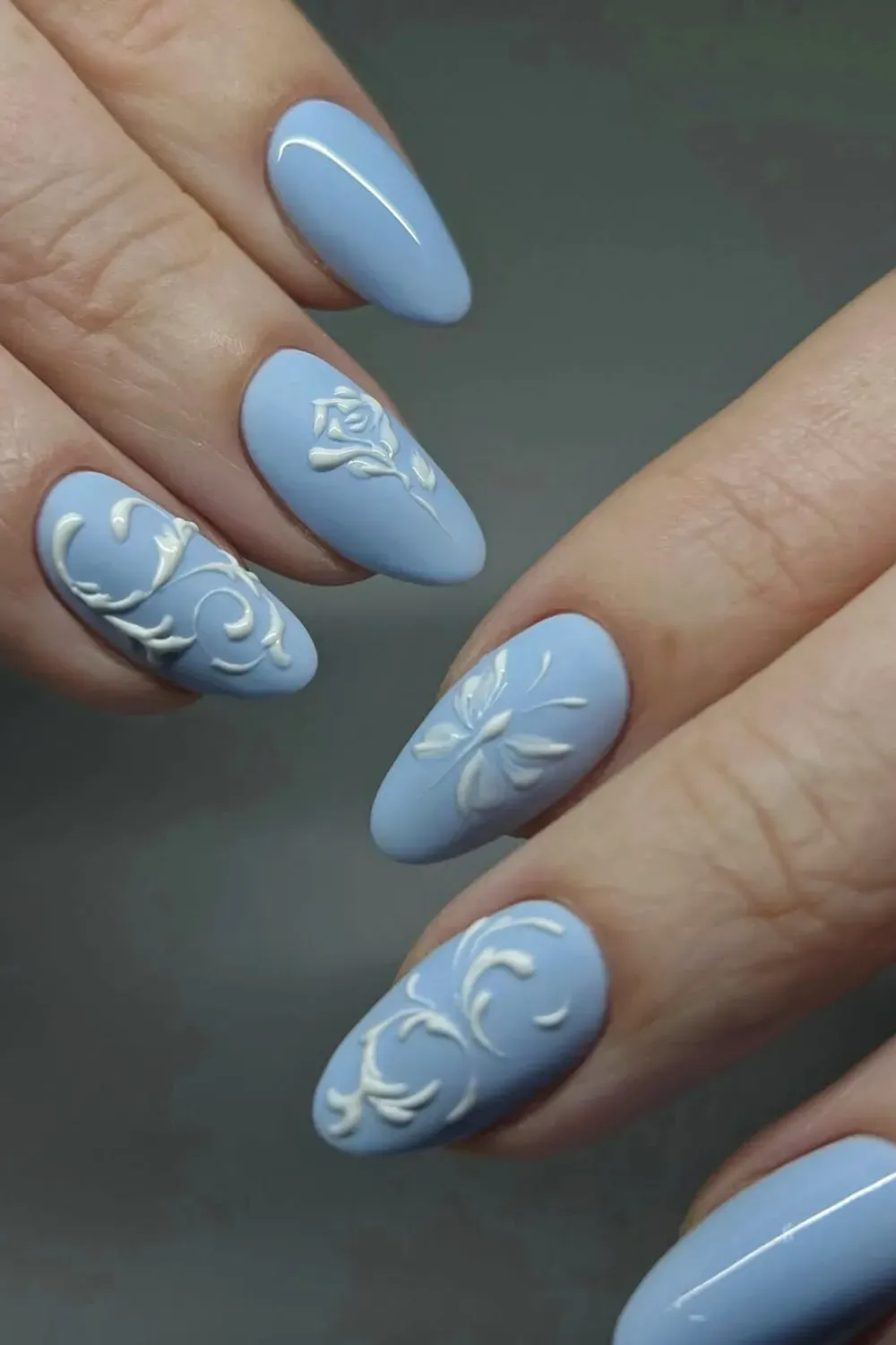 Close-up of light blue almond-shaped nails with white 3D embossed floral and swirl designs.