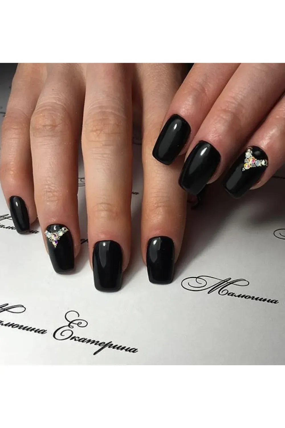 Square-shaped black glossy nails, with an accent nail featuring a cluster of iridescent rhinestones arranged in a triangle/heart shape.
