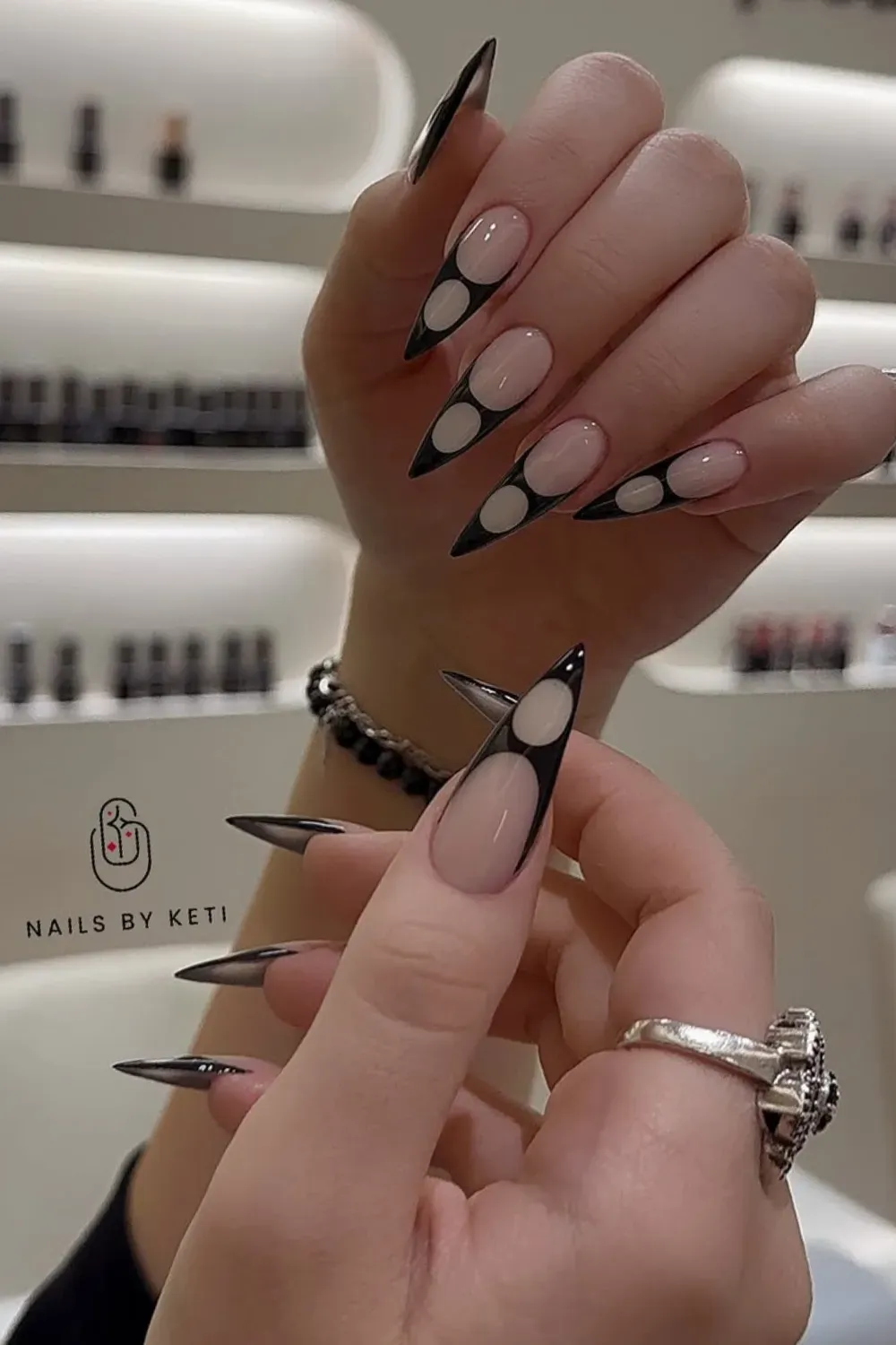 Hands showcasing very long, sharp stiletto nails with a nude base, bold black outlines, and circular geometric cut-out designs.