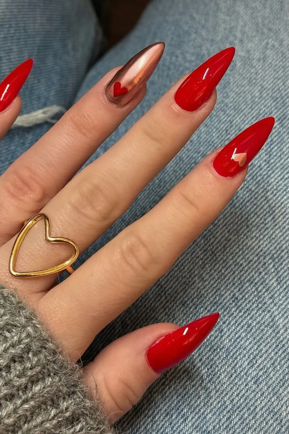 Hand with long red stiletto nails, one nail is metallic rose gold with a red heart, and others have small gold heart accents.