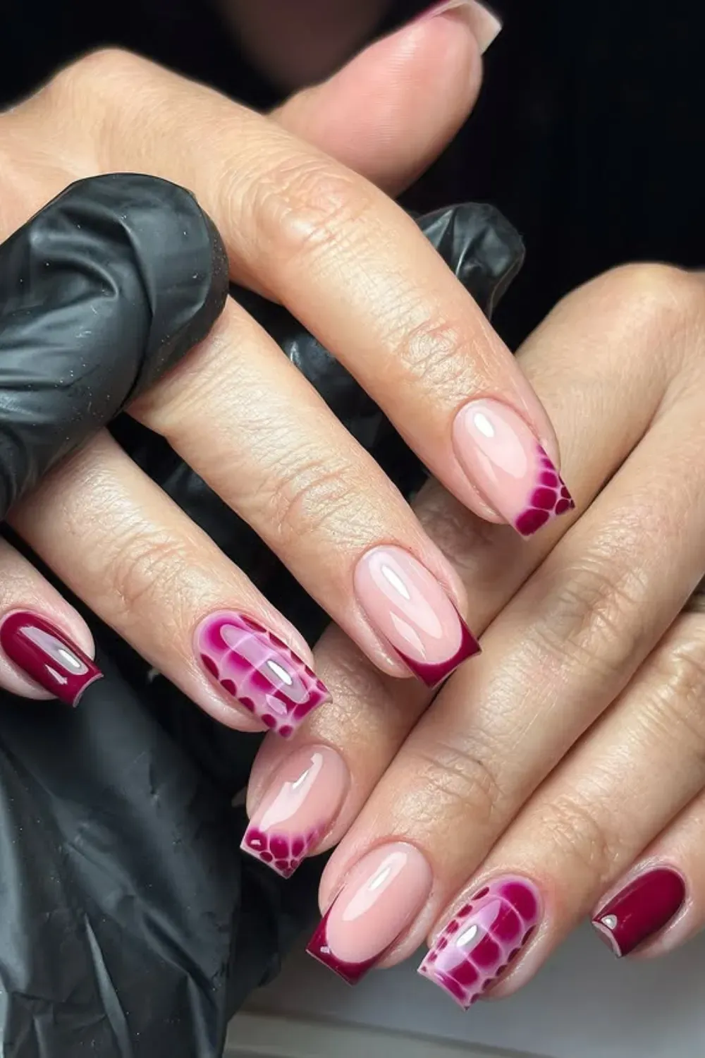 Hands with square-shaped nails featuring nude bases, burgundy French tips, and geometric web patterns in lighter red.