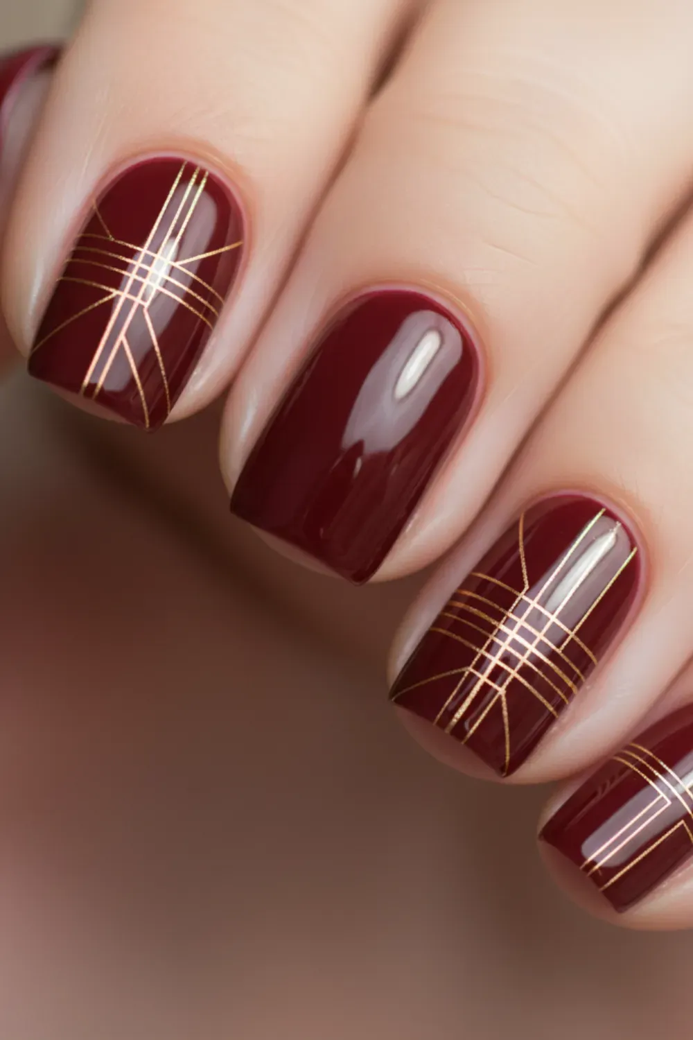 Close-up of a hand with glossy square burgundy nails, some adorned with intricate gold geometric grid patterns.