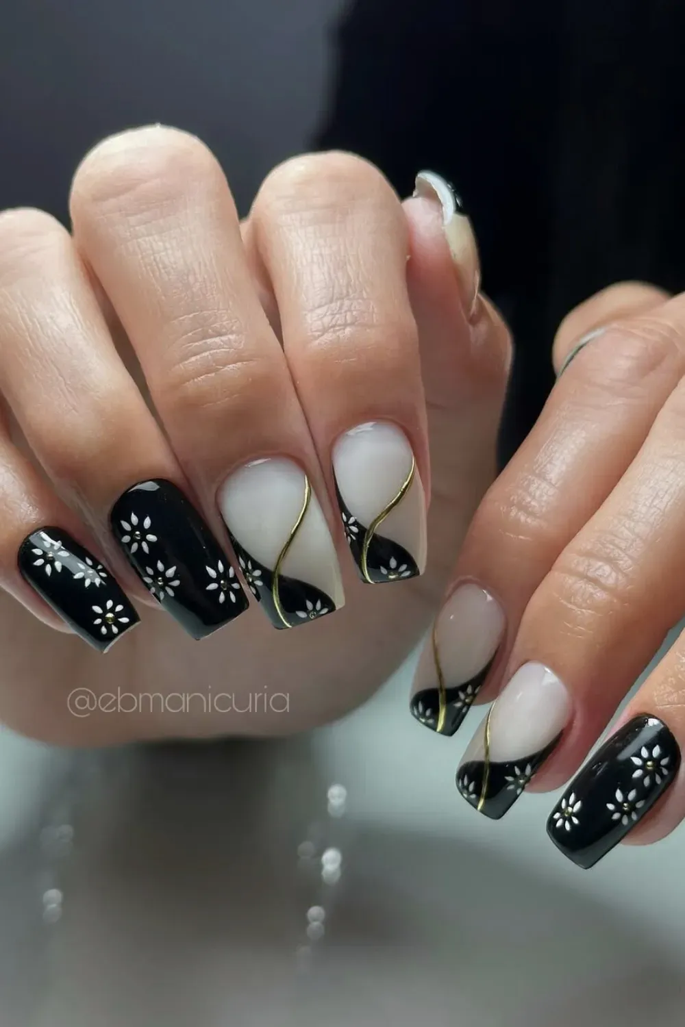 Square nails featuring a bold black and white design with delicate white flowers and curved gold metallic lines separating the colors.