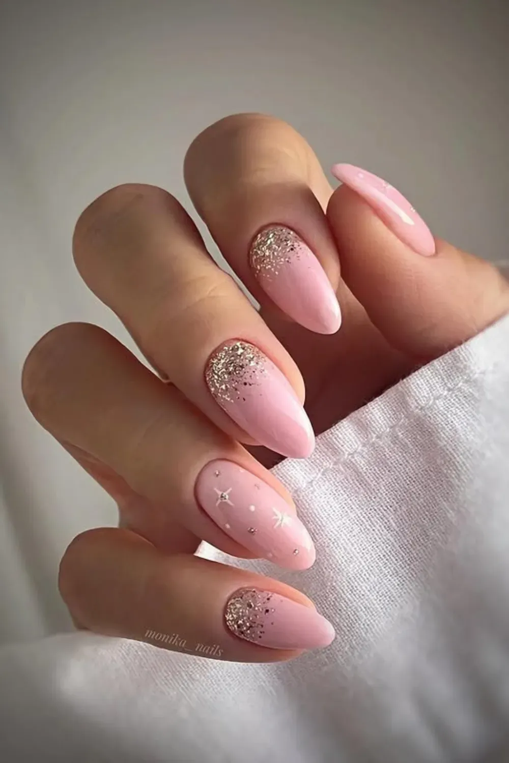 Hand with light pink almond-shaped nails; some have gold glitter at the cuticle, others have small white stars and dots.