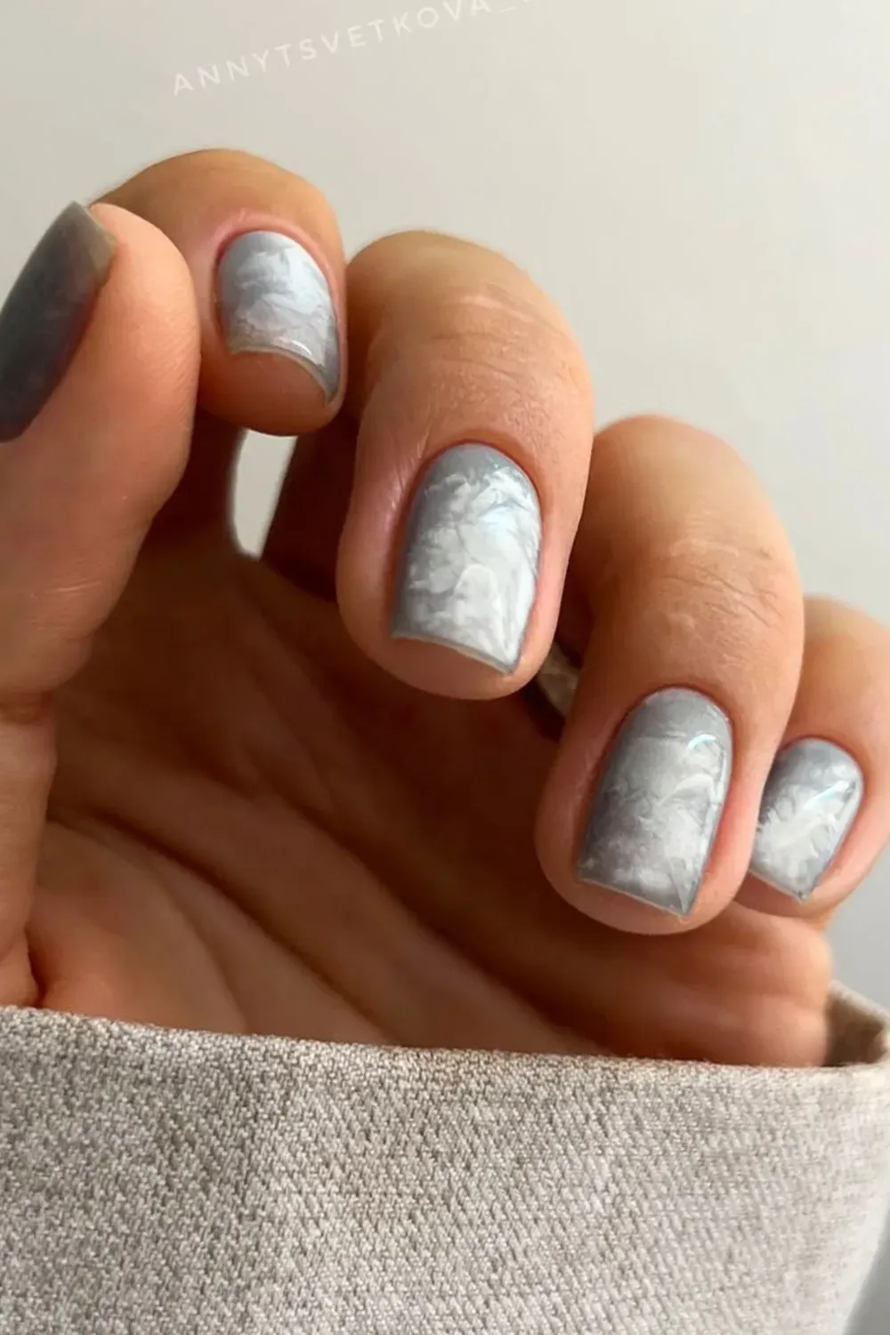 Hand with short square grey marble nails, resting on a light-colored sleeve.