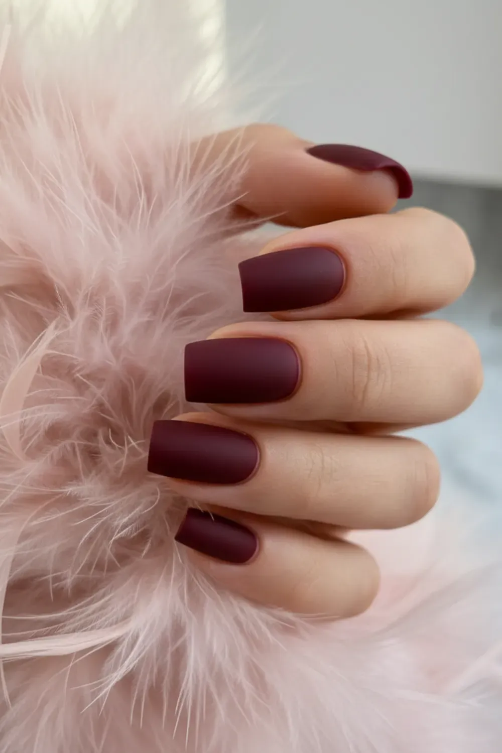Hand holding a pink feather, showing short square nails with a matte finish in a deep burgundy color.